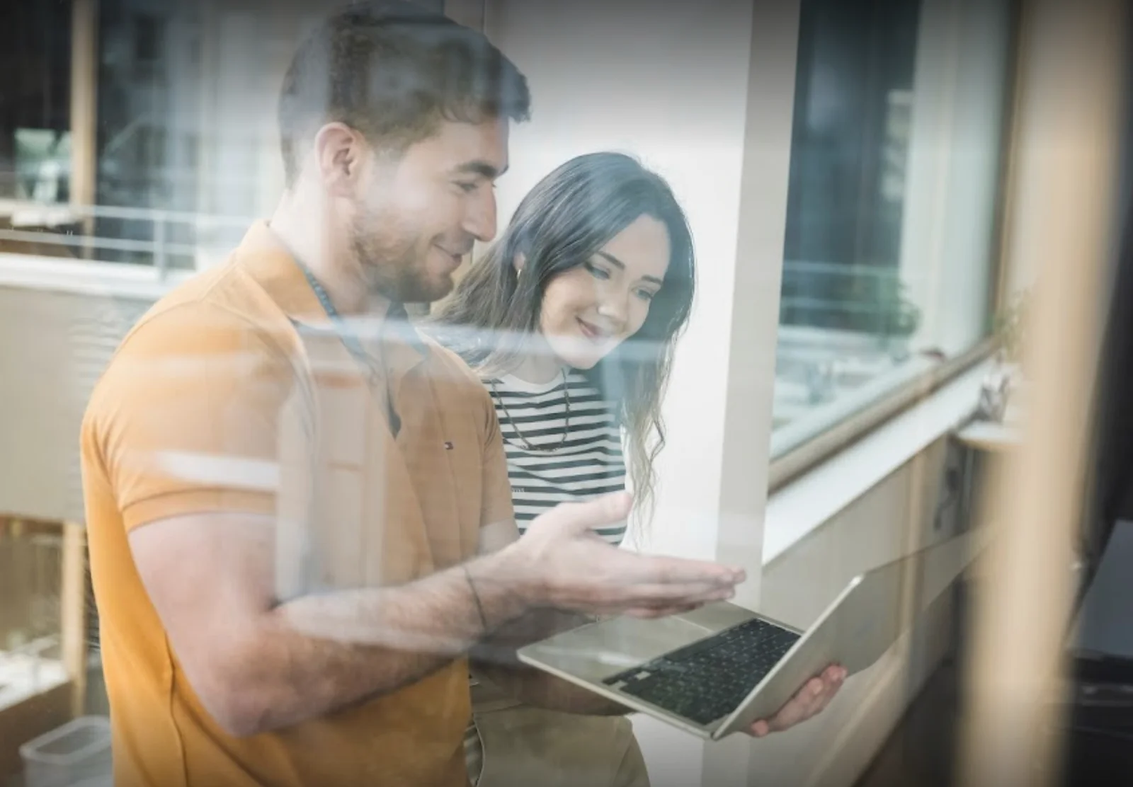 Zwei Personen stehen in einem Innenraum zusammen, lächeln und schauen auf einen Laptop-Bildschirm, während sie KI für Unternehmen erforschen, mit Spiegelungen auf einer Glasfläche vor ihnen. - digiak webdesign und entwicklung